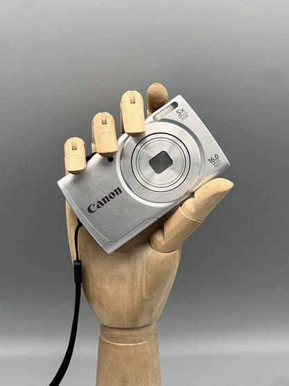 Silver Canon PowerShot A2500 with its lens closed and wrist strap attached, held in a wooden display hand