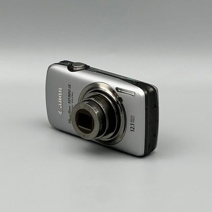 Angled front view of a silver Canon PowerShot SD980 IS showing its extended lens and black side panel