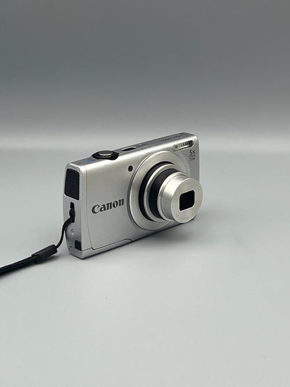 Angled front view of a silver Canon PowerShot A2500 showing its extended lens and wrist strap attachment