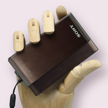 Brown Sony Cyber-shot DSC-T900 with its sliding lens cover closed, held in a wooden display hand