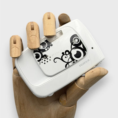 White Fujifilm FinePix Z30 with a black swirl pattern, held in a wooden display hand with its lens cover closed.