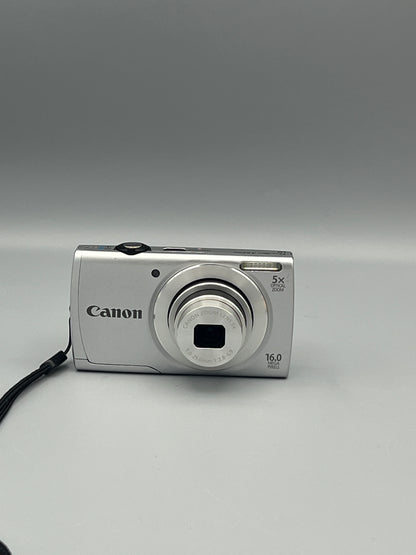 Front view of a silver Canon PowerShot A2500 with its lens extended and wrist strap to the side
