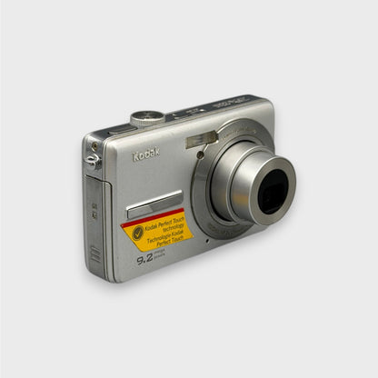 Angled front view of a silver Kodak EasyShare M320 with its lens extended and yellow feature sticker visible