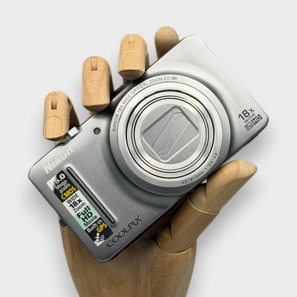 Silver Nikon Coolpix S9300 digital camera resting in a wooden hand with the lens closed