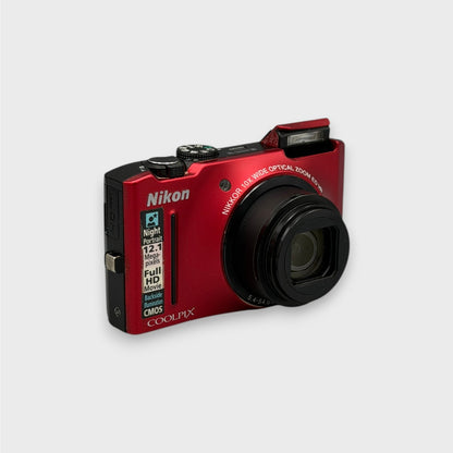 Angled front view of a red Nikon Coolpix S8100 with its lens extended and pop-up flash raised