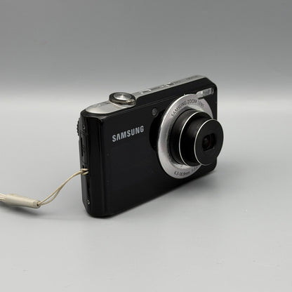 Angled front view of a black Samsung PL100 showing its extended lens and silver top panel