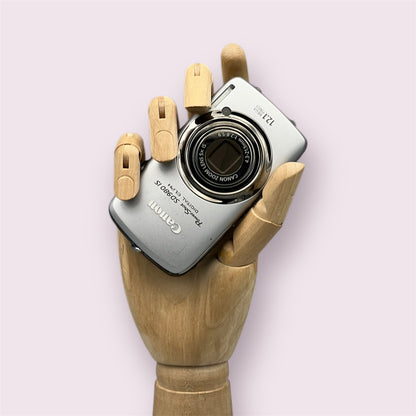 Silver Canon PowerShot SD980 IS with its lens closed, held in a wooden display hand