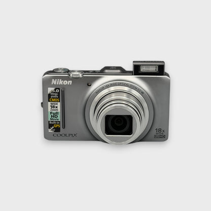 Front view of a silver Nikon Coolpix S9300 with its lens extended and flash raised