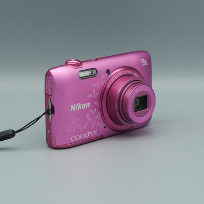 Angled front view of a pink floral Nikon Coolpix S3600 showing its extended lens and wrist strap attachment