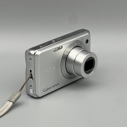 Angled front view of a silver Sony Cyber-shot DSC-W220 showing its extended lens and wrist strap attachment