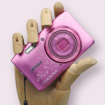 Pink Nikon Coolpix S3600 with a floral design and closed lens, held in a wooden display hand