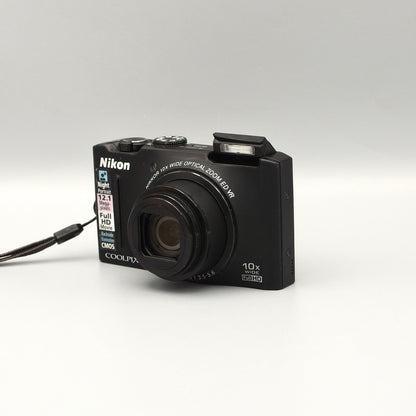 Black Nikon Coolpix S8100 viewed from a front angle showing the extended lens, raised flash and top controls.