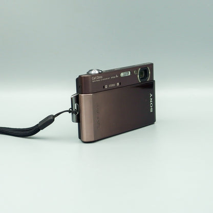 Angled front view of a brown Sony Cyber-shot DSC-T900 showing its open lens cover and wrist strap attachment