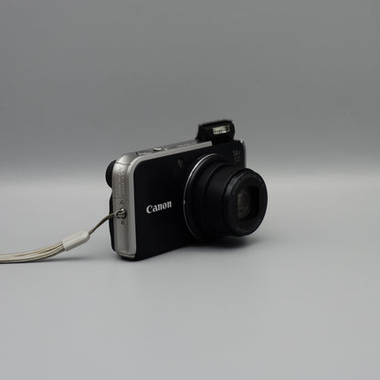 Angled front view of a black Canon PowerShot SX210 IS with its lens extended and flash raised