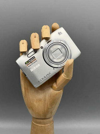 Silver Nikon Coolpix S3600 with its lens closed, held in a wooden display hand