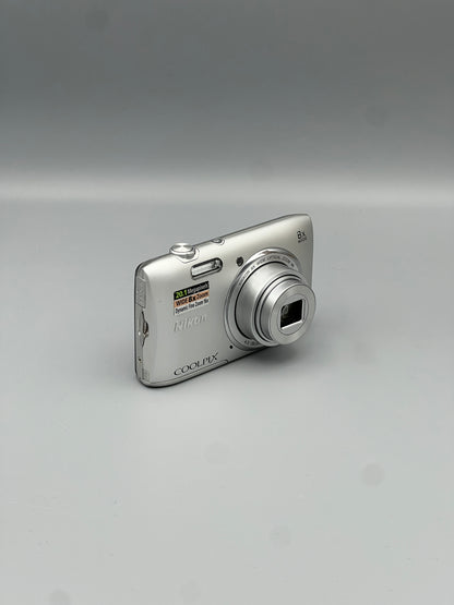 Angled front view of a silver Nikon Coolpix S3600 showing its extended lens and side connection cover