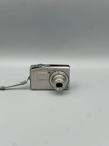 Silver Fujifilm FinePix F50FD front view with its lens extended and wrist strap attached.