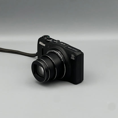 Black Nikon Coolpix S9700 viewed from above at a front angle with its zoom lens extended.
