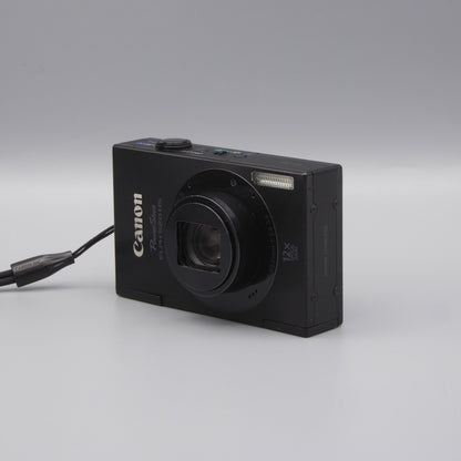 Black Canon PowerShot ELPH 520 HS viewed from a front angle with its lens extended and wrist strap beside it.