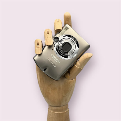 Silver Canon PowerShot SD900 with its lens closed, held in a wooden display hand