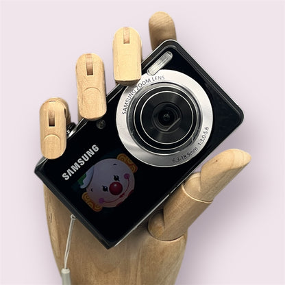 Black Samsung TL205 digital camera in a wooden hand with the lens open and a clown graphic on the front display