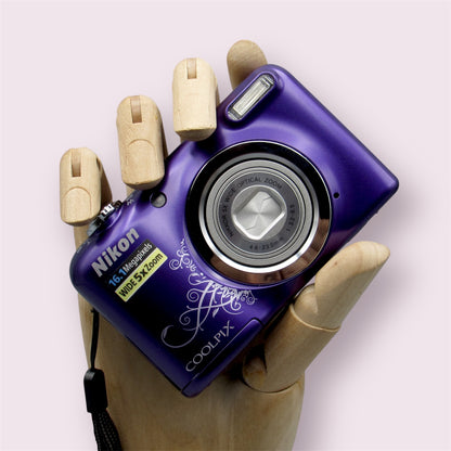 Purple Nikon Coolpix A10 with a decorative swirl pattern, held in a wooden display hand with its lens closed.