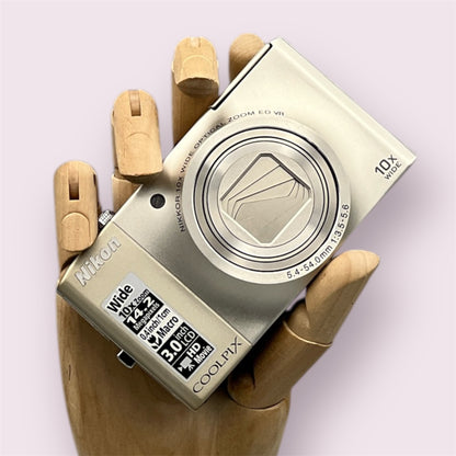 Silver Nikon Coolpix S8000 with its lens closed, held in a wooden display hand