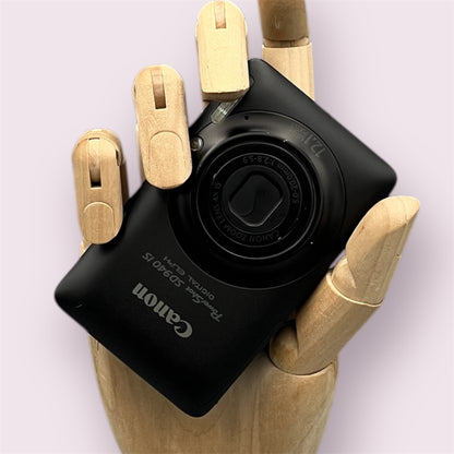 Black Canon PowerShot SD940 IS held in a wooden display hand with its lens closed.