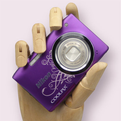 Purple Nikon Coolpix A100 digital camera in a wooden hand with a white decorative pattern and closed lens