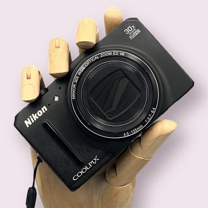 Black Nikon Coolpix S9700 held in a wooden display hand with its lens closed.