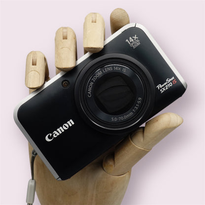 Black Canon PowerShot SX210 IS digital camera resting in a wooden hand with the lens closed