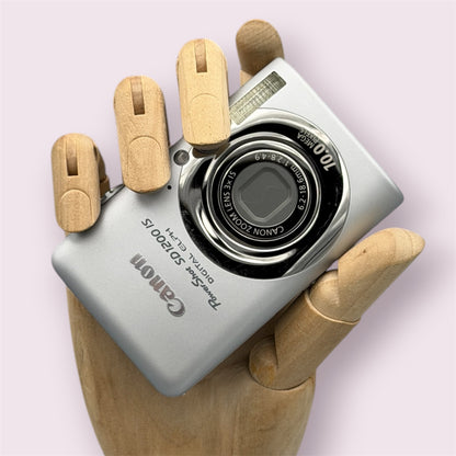 Silver Canon PowerShot SD1200 IS with its lens closed, held in a wooden display hand