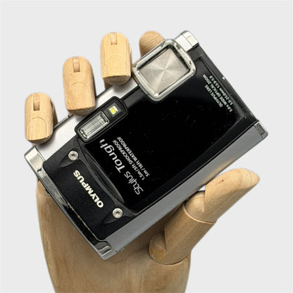 Black and silver Olympus Tough 6020 digital camera resting in a wooden hand with the front visible