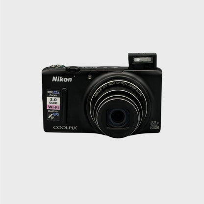 Front view of a black Nikon Coolpix S9500 with its lens extended and flash raised