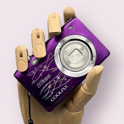 Purple Nikon Coolpix S2800 with a decorative line pattern, held in a wooden display hand with its lens closed.