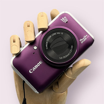 Purple Canon PowerShot SX220 HS digital camera resting in a wooden hand with the lens closed
