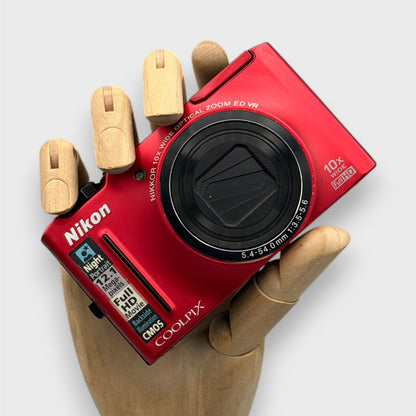 Red Nikon Coolpix S8100 with its lens closed, held in a wooden display hand