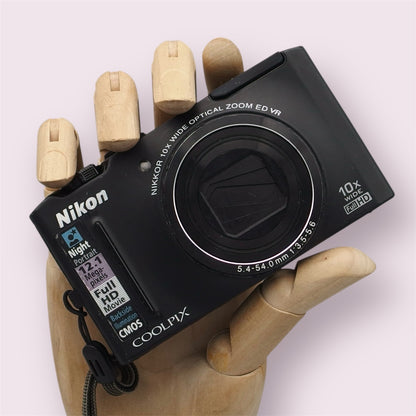 Black Nikon Coolpix S8100 held in a wooden display hand with its lens closed.