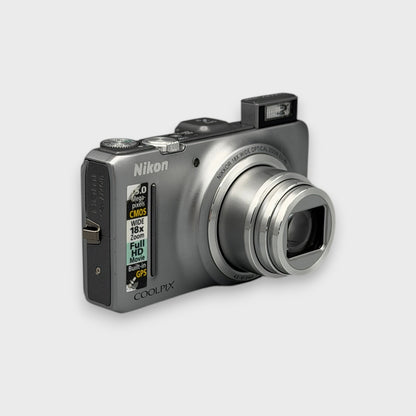 Angled front view of a silver Nikon Coolpix S9300 with its lens extended and flash raised