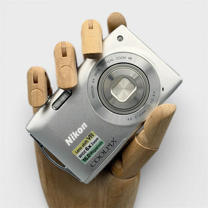 Silver Nikon Coolpix S3300 digital camera resting in a wooden hand with the lens closed