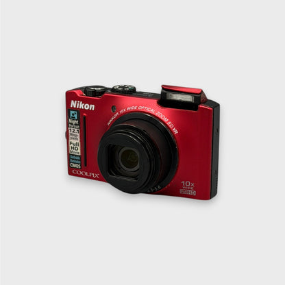 Red Nikon Coolpix S8100 viewed from the opposite front corner with its lens extended and flash raised