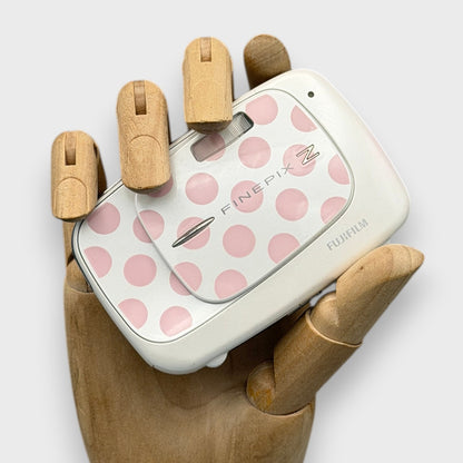 White Fujifilm FinePix Z37 with pink polka dots, held in a wooden display hand with its lens cover closed.