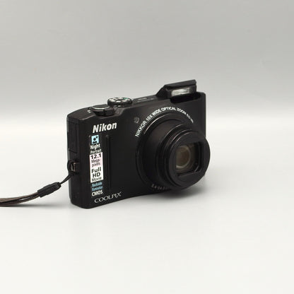 Black Nikon Coolpix S8100 viewed from a front angle with its lens extended and pop-up flash raised.